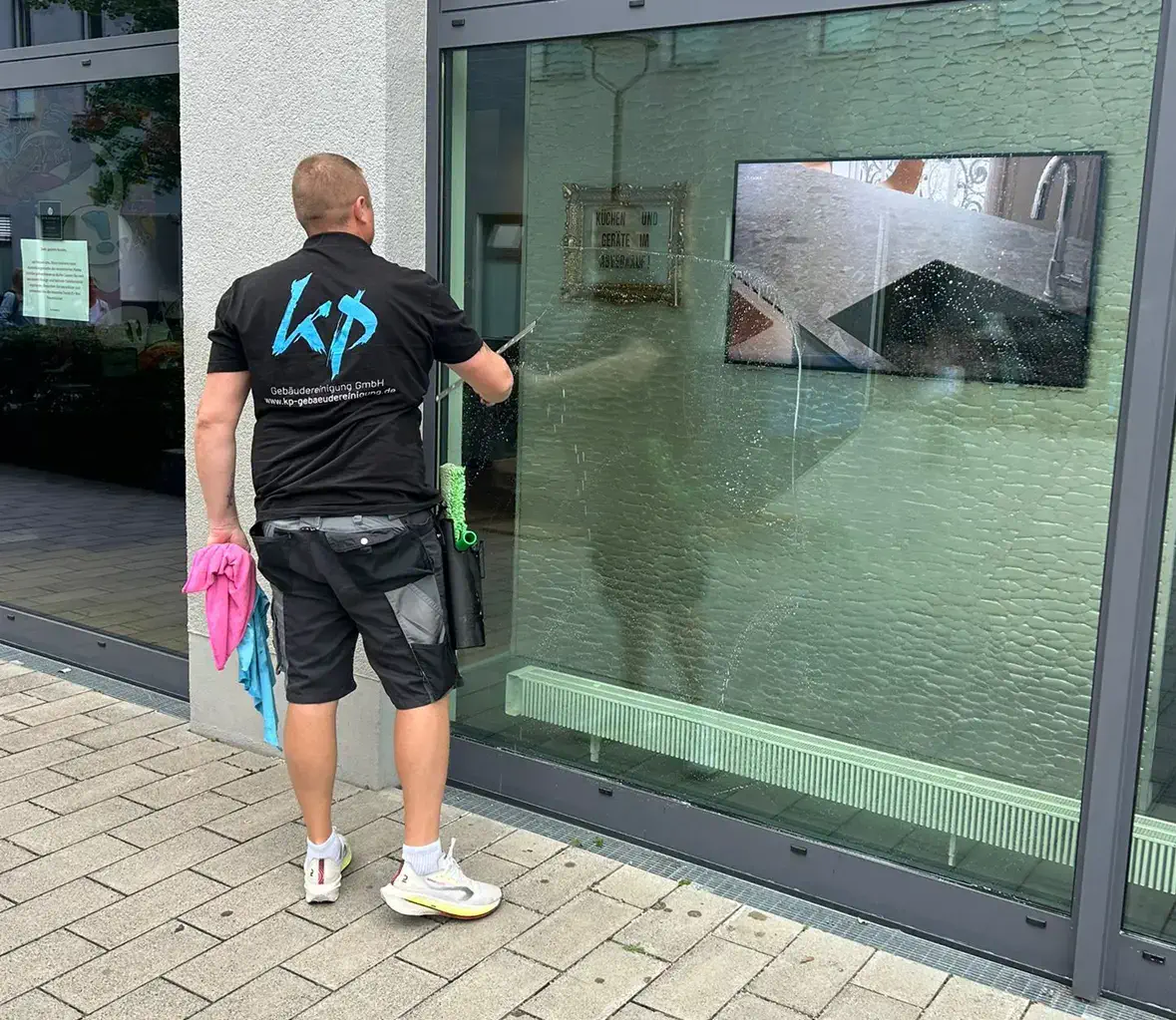 Ein Mitarbeiter der KP Gebäudereinigung GmbH aus München reinigt eine große Schaufensterscheibe. Der Mann trägt ein schwarzes T-Shirt mit dem blauen Logo der Firma und eine kurze, schwarze Arbeitshose. In der linken Hand hält er ein pinkes Reinigungstuch, in der rechten Hand einen Fensterschaber.